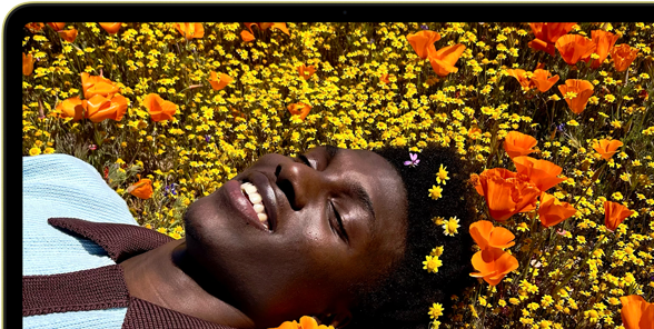 MacBook Neo, citrus color, screen shows a smiling person lying in a sunny field of flowers