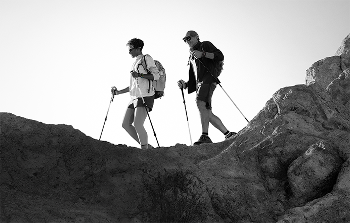 Two people wearing Apple Watch devices and hiking on a rocky trail with poles and gear