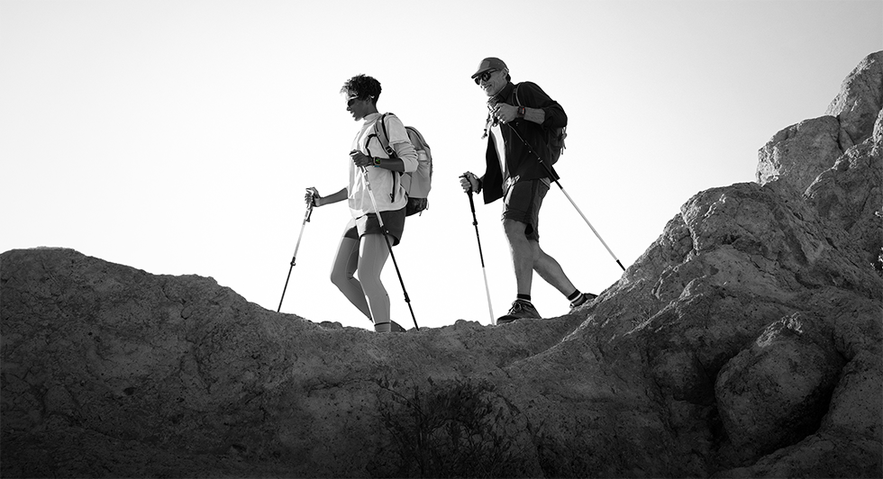 Two people wearing Apple Watch devices and hiking on a rocky trail with poles and gear